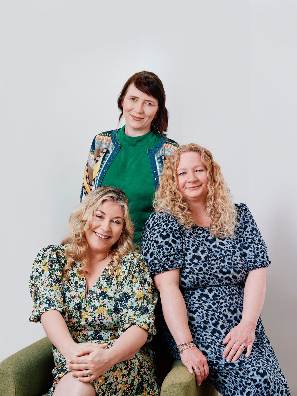 Three smiling women with curly hair posing together against a light gray background, two seated and one standing behind.