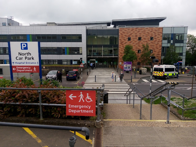 Entrance to a hospital building with signs directing to the North Car Park and Emergency Department, pedestrians using a crosswalk, and a parked ambulance nearby.